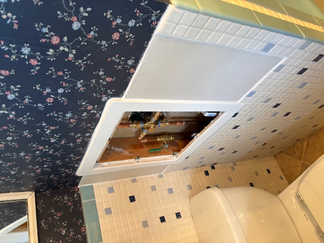 Bathroom plumbing access panel in tiled wall, featuring floral wallpaper and adjacent toilet, highlighting renovation needs for effective bathroom remodeling.